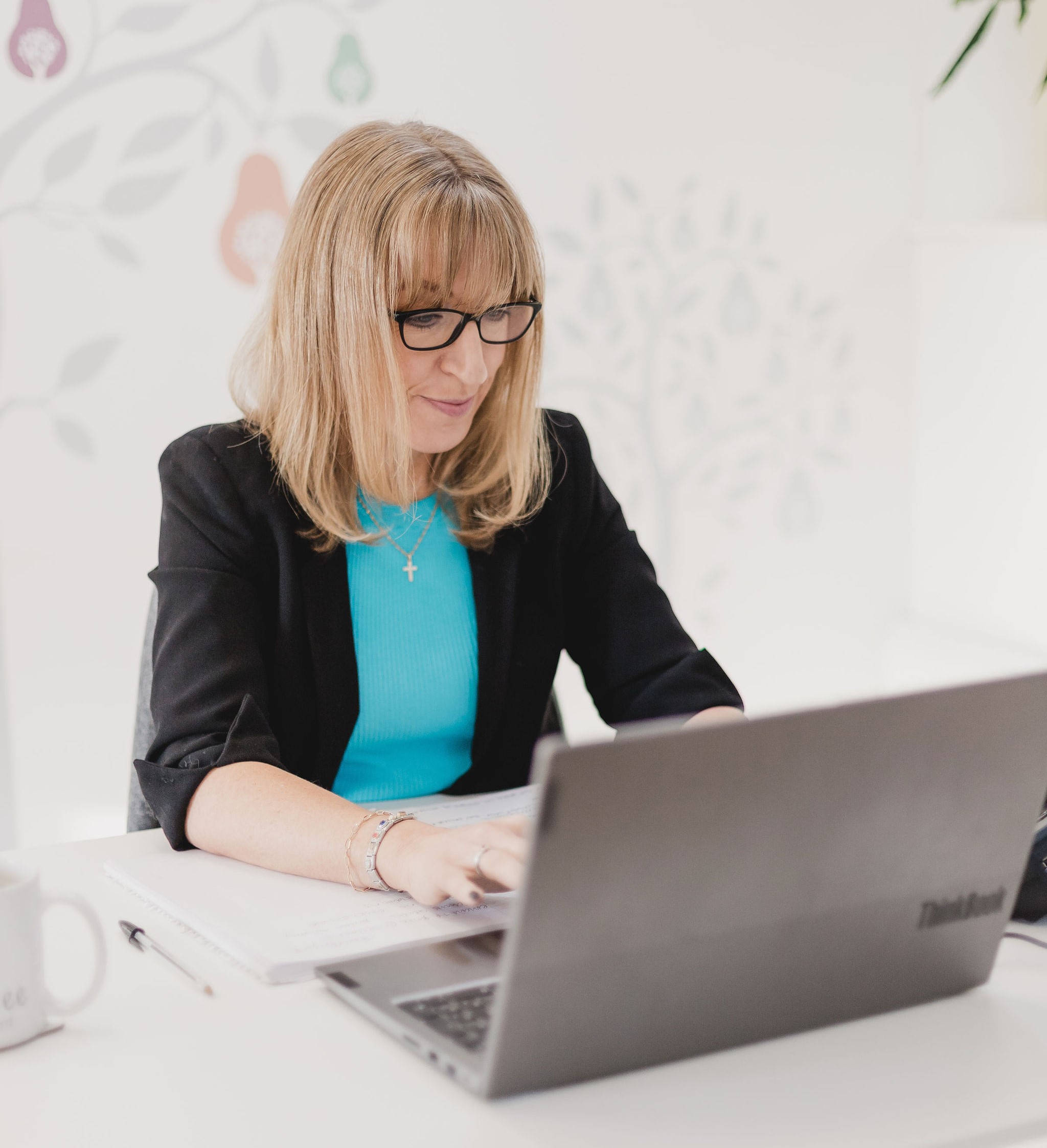 Photo of Kate at her desk in front of a colourful Pear Tree Recruitment logo and working on her laptop